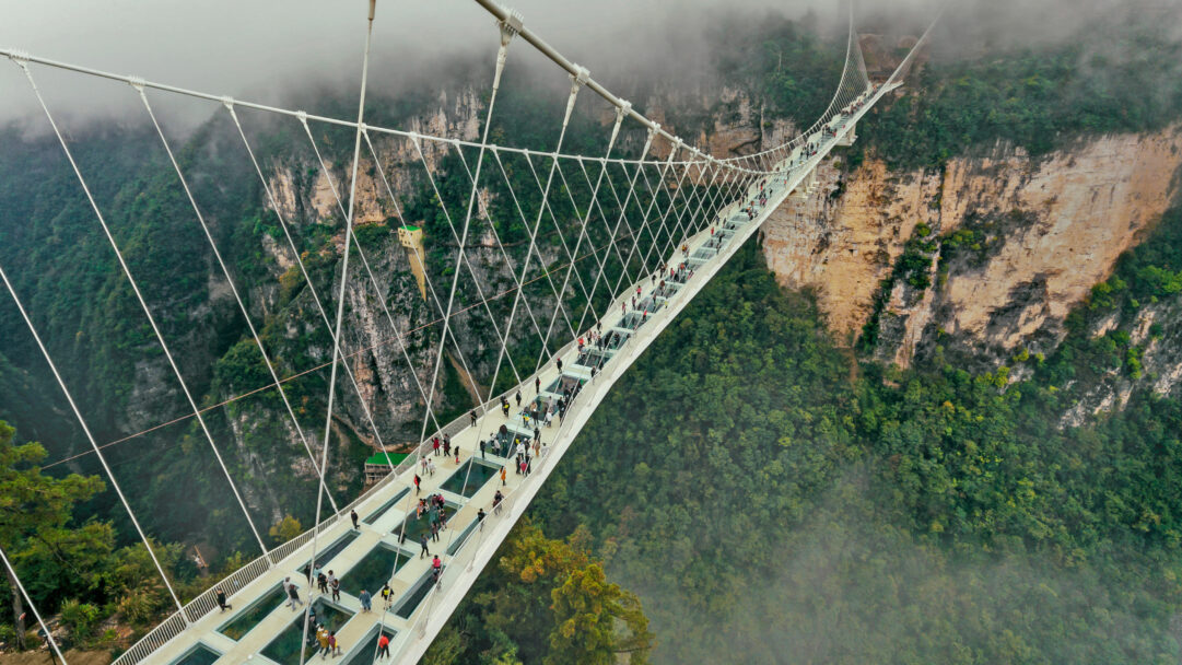 A breathtaking 4K wallpaper depicting the Zhangjiajie Glass Bridge in Hunan, China, stretching across a deep mountain gorge on a foggy day. Numerous visitors traverse the transparent sections of the bridge, offering a thrilling view down to the lush, mist-shrouded peaks and valleys below, emphasizing the bridge's daring design amidst the dramatic natural landscape.