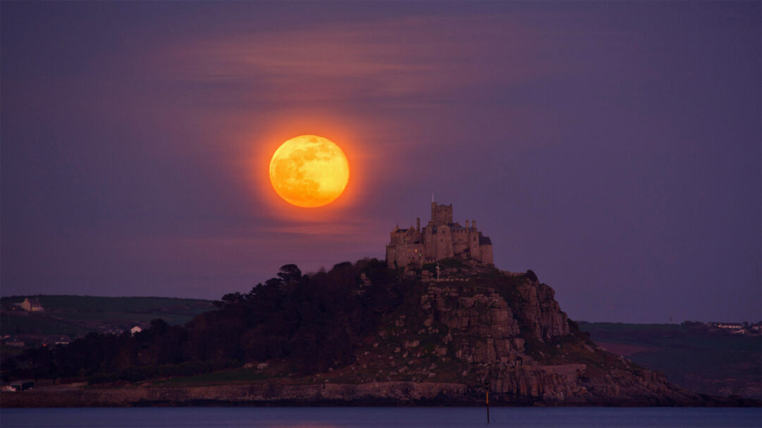 An ethereal 4K wallpaper showcasing a majestic island castle perched on a rocky outcrop, illuminated by the radiant 2017 April Pink Full Moon. The intensely bright, amber-hued full moon dominates the purplish-pink twilight sky, casting a serene glow that reflects softly on the calm water below.