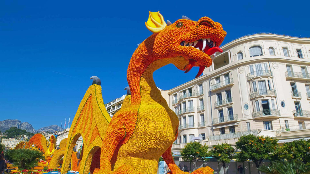An astonishing 4K wallpaper presents a colossal citrus dragon sculpture from the 2019 Menton Lemon Festival in France, prominently displayed against a clear blue sky and ornate buildings. The intricate texture of thousands of yellow lemons and orange oranges forming the dragon's scales radiates a vibrant, festive energy under the brilliant sunshine.