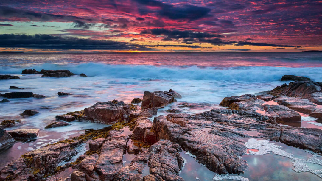 A captivating 4K wallpaper showcases Acadia National Park in Maine, featuring a dramatic ocean sunset over its distinctive rocky shore. Vibrant crimson and purple clouds adorn the sky as frothy waves crash against the textured dark rocks, reflecting the fading light in tranquil tide pools.