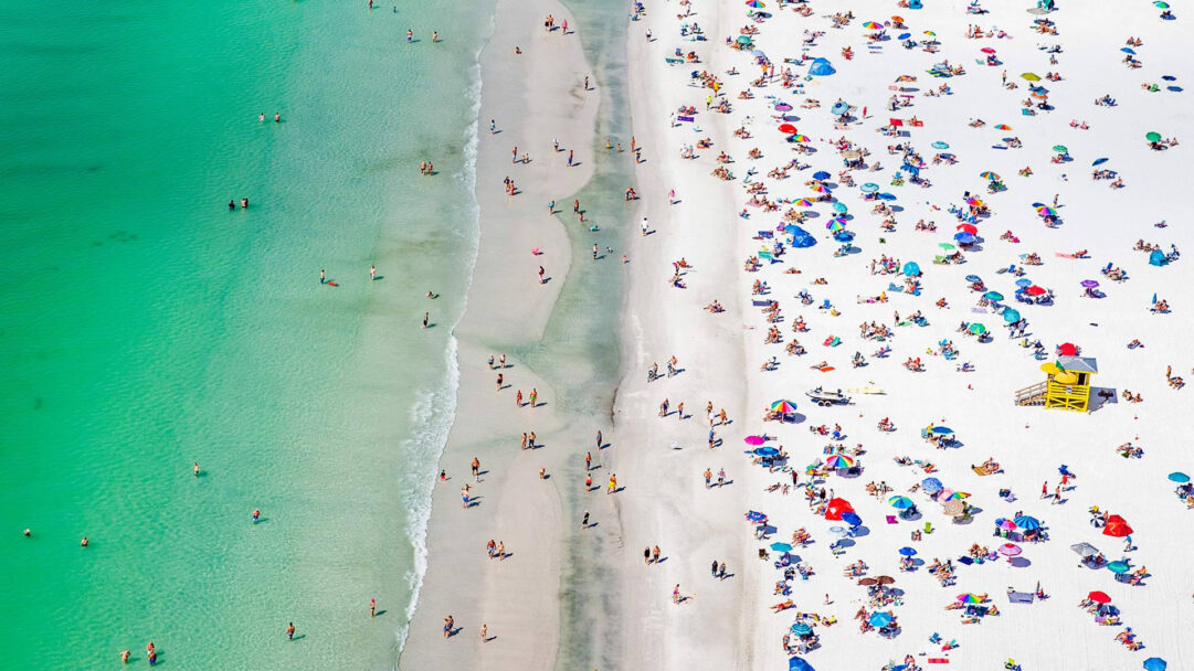 An expansive 4K wallpaper showcases an aerial view of the crowded Siesta Key Florida shoreline, teeming with beachgoers on pristine white sand and in turquoise water. The vibrant interplay of countless colorful umbrellas and tiny figures against the clear, inviting green-blue ocean defines a lively, joyful summer atmosphere.