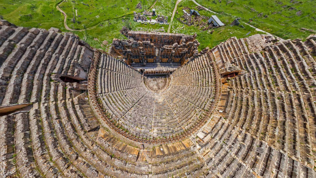 An awe-inspiring 4K wallpaper presenting an aerial view of the remarkably preserved Ancient Theater of Hierapolis, nestled within a sprawling green archaeological landscape. The concentric, weathered stone seating tiers create a dramatic, sun-drenched semi-circular form, contrasting sharply with the vibrant green terrain surrounding the ancient site.