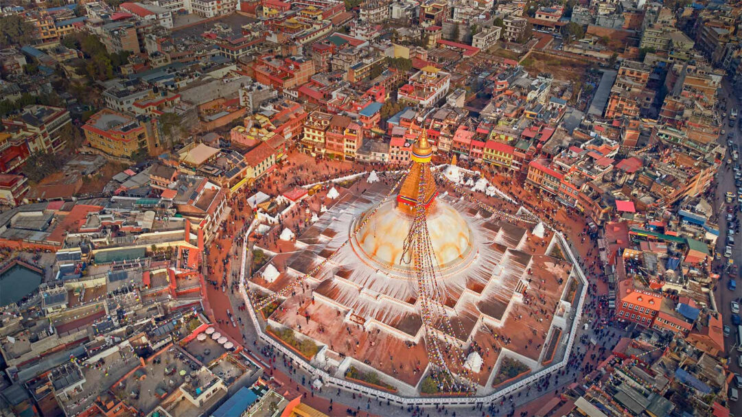 A magnificent 4K wallpaper displaying an aerial view of the grand Boudhanath Stupa in Kathmandu, Nepal, encircled by a bustling urban landscape of densely packed buildings and streets. Golden sunlight illuminates the stupa's majestic white dome and golden spire, adorned with colorful prayer flags, as numerous people gather and walk around its multi-tiered base, conveying a sense of vibrant activity and spiritual reverence.
