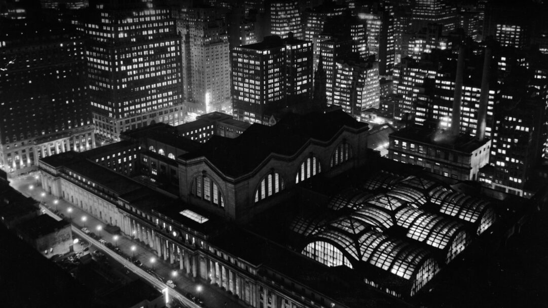An evocative 4K wallpaper capturing an aerial view of the historic Penn Station amidst the sprawling New York City skyline at night. The striking black and white imagery emphasizes the dramatic glow of countless illuminated windows across surrounding skyscrapers, creating a timeless and bustling urban atmosphere.
