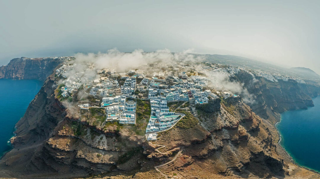 A captivating 4K wallpaper presents an aerial view of a picturesque cliffside town in Santorini, Greece, nestled precariously above the deep blue Aegean Sea. Wisps of soft fog drift through the iconic white buildings, creating a mystical and serene atmosphere against the rugged volcanic landscape.