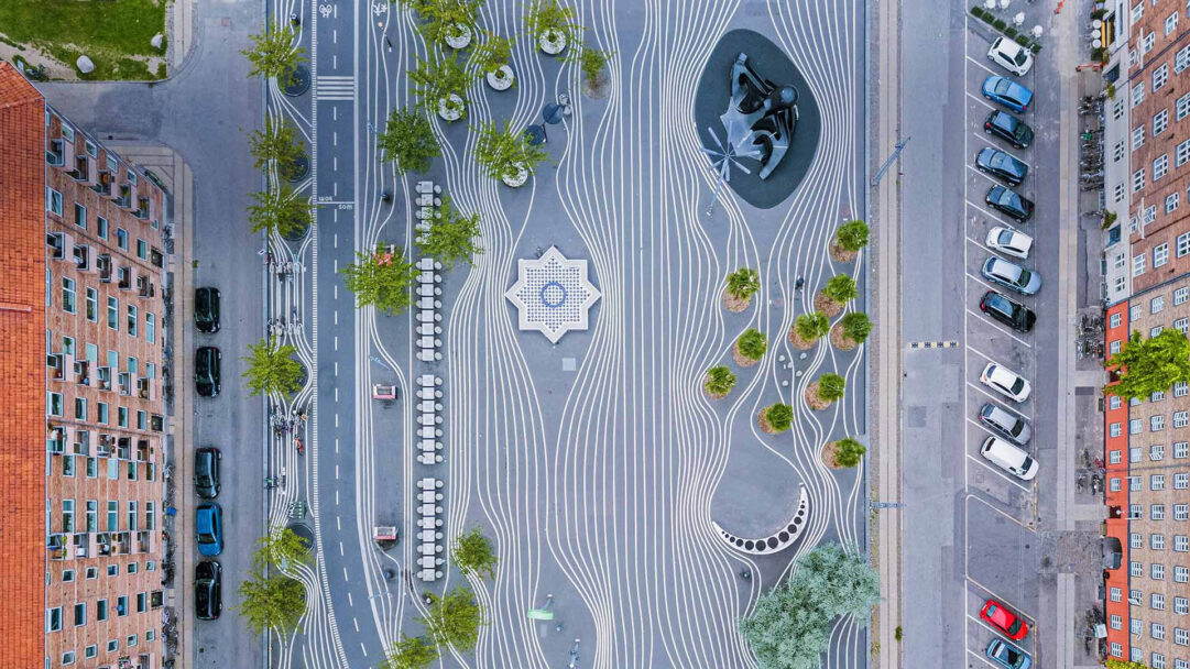 A masterful 4K wallpaper capturing an aerial view of Superkilen Park in Copenhagen, Denmark, showcasing its innovative urban design layout amidst surrounding city blocks. The park's striking visual elements include a vibrant interplay of flowing white lines on grey pavement, interspersed with diverse international urban features such as a star-shaped fountain, a dark abstract sculpture, and various green planting zones.