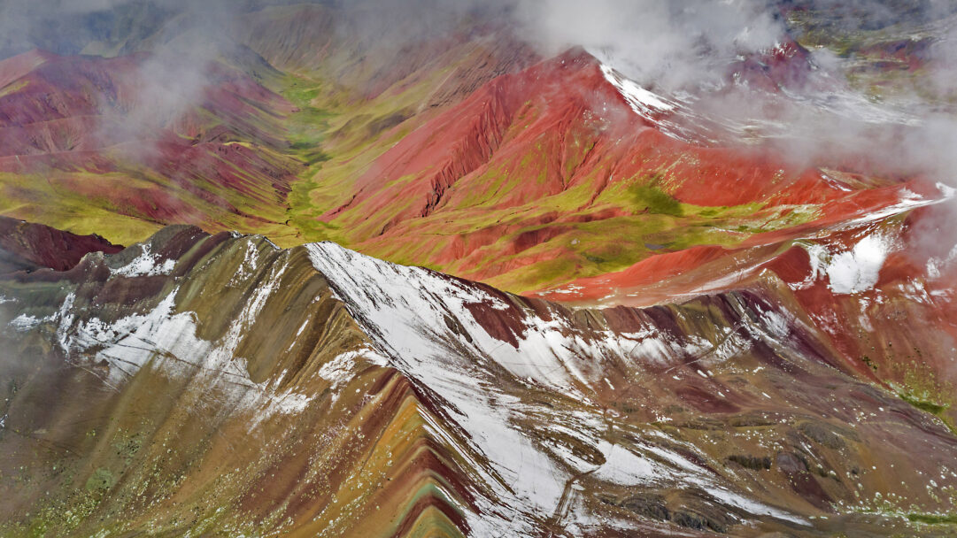 An awe-inspiring 4K wallpaper showcasing an aerial view of Vinicunca, Peru, revealing its iconic colorful mountains and dramatic snowy peaks. The vibrant geological striations of red, green, and brown create a breathtaking tapestry, accentuated by pristine white snow and ethereal clouds drifting through the high-altitude landscape.