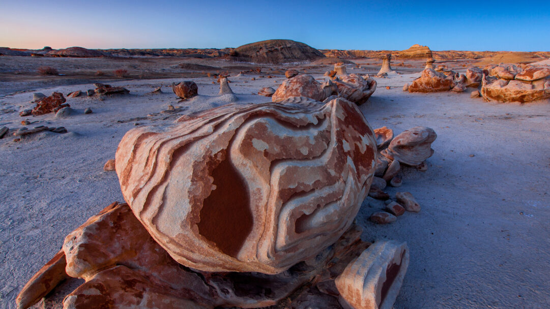 A mystical 4K wallpaper revealing the 'Alien Egg Hatchery' formations nestled within the Bisti De-Na-Zin Wilderness desert landscape. The striking wavy patterns of ochre and white on the large, egg-like rock in the foreground contrast with the cool, sandy ground and pale blue sky, creating a serene yet enigmatic atmosphere.