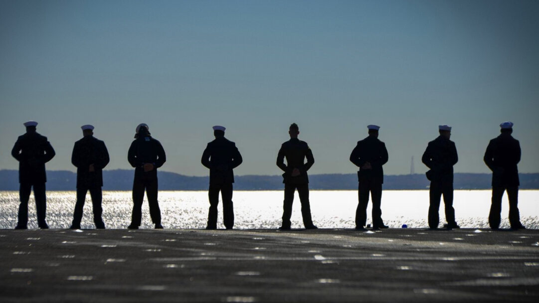 A majestic 4K wallpaper showcasing eight Amphibious Assault Ship USS Iwo Jima sailors standing in silhouette on the deck, overlooking the Hudson River. The golden shimmer of the river, backlighting their stoic forms and crisp white sailor caps against the vast expanse of the sky, creates a powerful and contemplative scene.