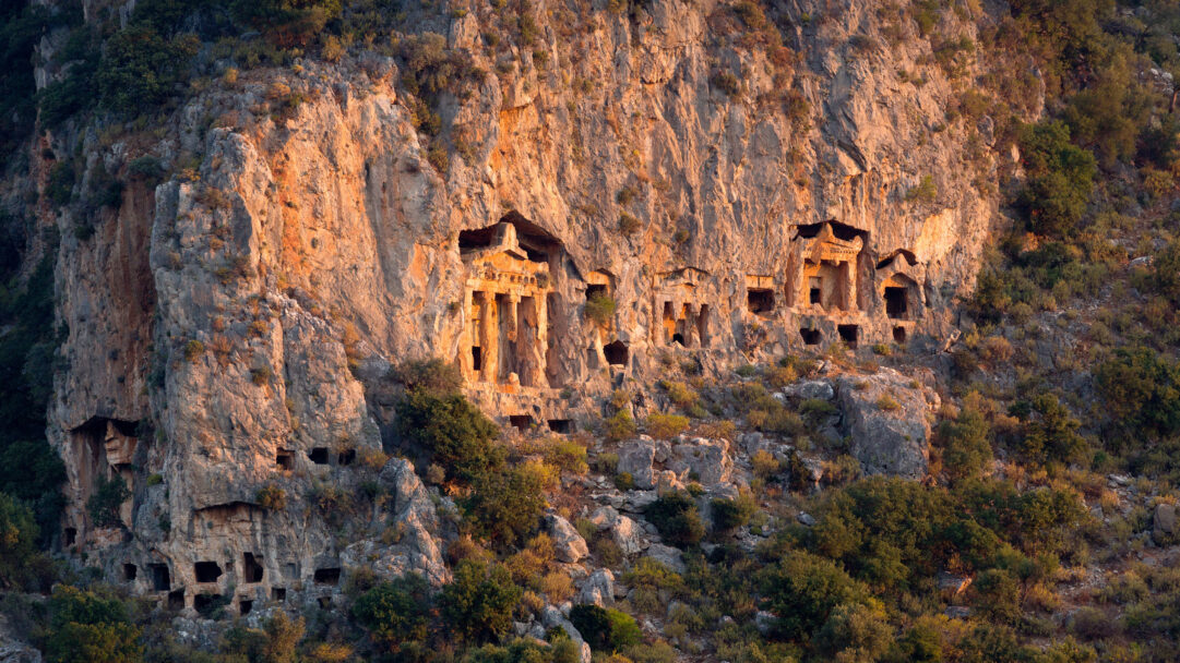 An awe-inspiring 4K wallpaper showcasing ancient Lycian rock tombs intricately carved into the towering Dalyan Cliff face. The warm, golden light of sunset bathes the cliff, highlighting the ornate facades of the tombs and casting dramatic shadows that emphasize their age and grandeur.