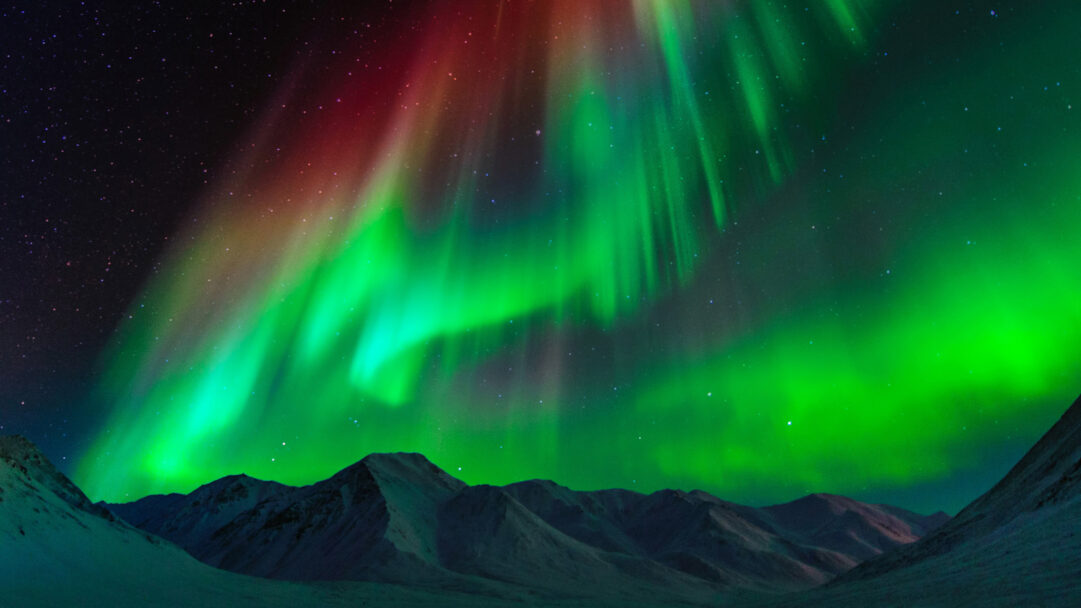 A breathtaking 4K wallpaper capturing the vibrant Aurora Borealis dancing across the night sky above the majestic Brooks Range in Alaska. Its intense green and crimson ribbons dramatically streak through the darkness, illuminating the snow-capped mountains below with an ethereal glow.