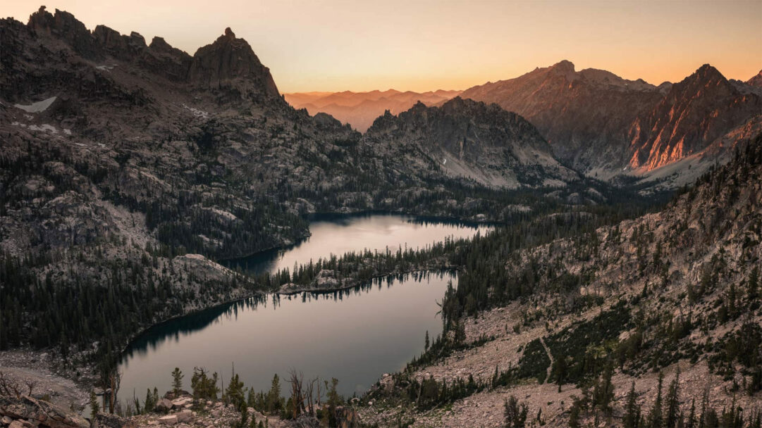 A breathtaking 4K wallpaper showcasing Baron Lake and Upper Baron Lake nestled within the rugged Sawtooth Wilderness at sunset. The fading light bathes the distant jagged peaks in warm orange hues, reflecting softly on the dark, still waters of the lakes amidst the dense pine forests.