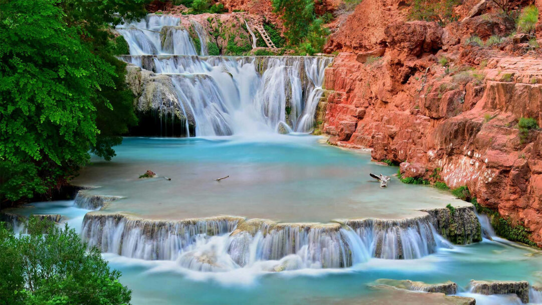 A breathtaking 4K wallpaper showcasing Beaver Falls, a multi-tiered waterfall cascading down into vivid blue-green pools within Havasu Creek in the Grand Canyon. The unique travertine formations create serene, shallow pools, while the vibrant turquoise water vividly contrasts with the rich red canyon walls and lush green foliage, evoking a tranquil and majestic natural beauty.