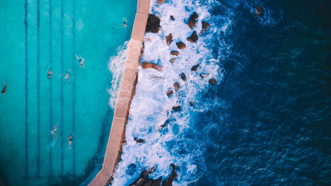 A captivating 4K wallpaper showcases the Bronte Baths ocean pool in Sydney from an aerial perspective, with swimmers enjoying the vibrant turquoise water. Waves crash dramatically against the pool's concrete edge and rugged rocks, creating a striking contrast between the tranquil, clear pool and the deep, foamy blue of the open ocean.