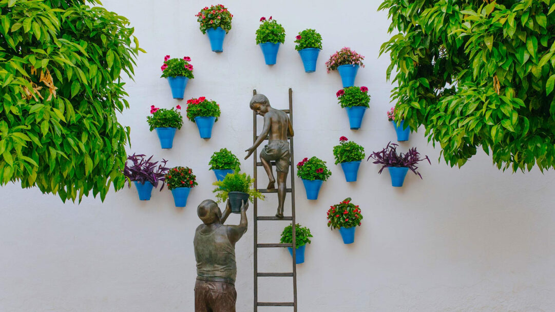 A charming 4K wallpaper showcases a bronze sculpture of a grandfather and child collaborating to care for an array of colorful potted plants mounted on a bright white wall. The detailed bronze figures, with the child on a ladder reaching for a plant passed by the grandfather, beautifully stand out against the vivid blues of the pots and the lush greens and vibrant reds of the diverse foliage.
