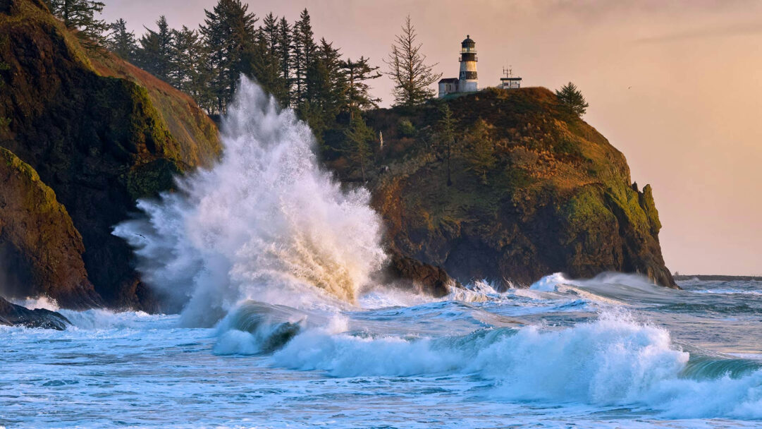 A dramatic 4K wallpaper showcasing the Cape Disappointment Lighthouse perched atop a rugged cliff overlooking the Washington coast near Ilwaco. Gigantic waves crash violently against the shore, with one colossal wave erupting in a dazzling spray bathed in the warm glow of the setting sun.