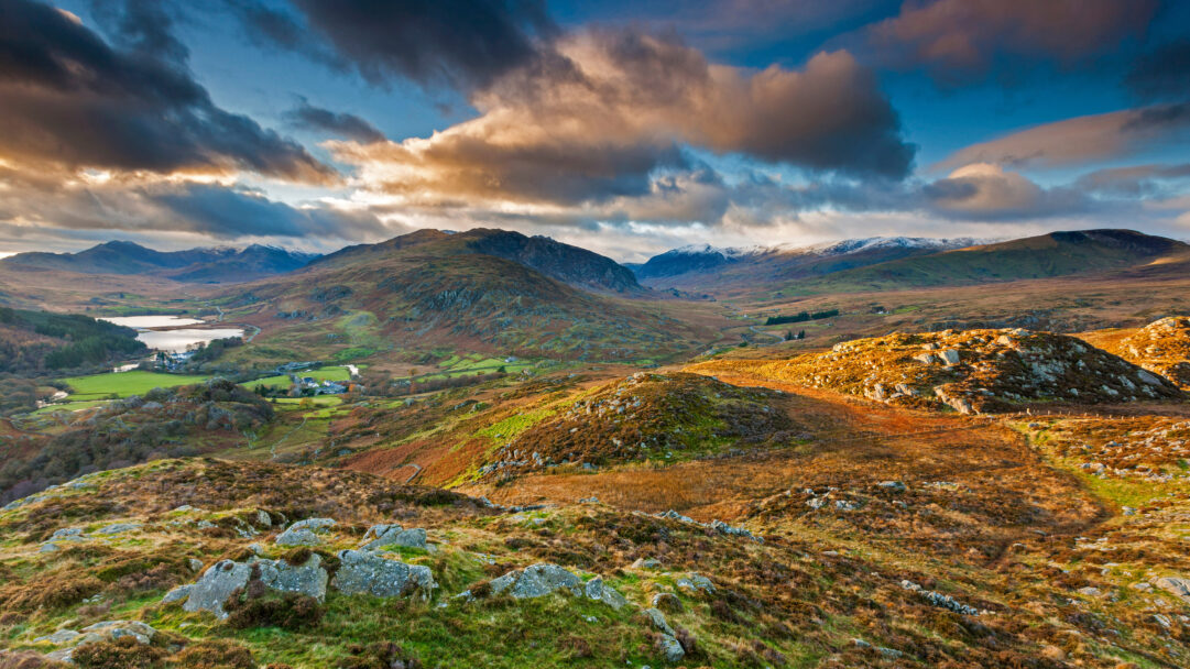An expansive 4K wallpaper captures the dramatic Capel Curig landscape within Snowdonia National Park, featuring a serene lake nestled amongst rolling mountains. Golden sunlight spectacularly illuminates the rugged, heather-covered foreground and the dramatic clouds, contrasting with the deep blue sky and accentuating the vast, wild beauty of the scene.