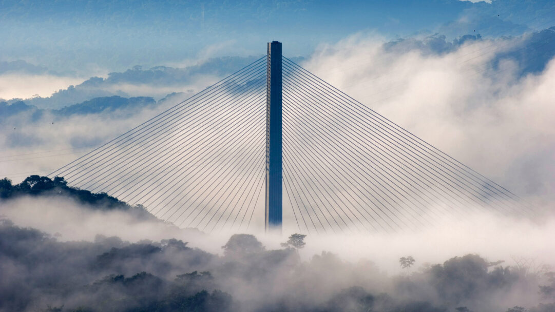 A breathtaking 4K wallpaper showcases the Centennial Bridge majestically towering above the dense, foggy canopy of Soberanía National Park. The bridge's central tower and radiating cables pierce through a sea of white mist, creating a dramatic contrast against the clearer blue sky and distant, hazy jungle peaks.