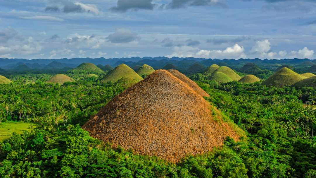 A spectacular 4K wallpaper featuring the unique Chocolate Hills of Bohol, Philippines, spread across a vast, lush landscape under a cloudy sky. The prominent central hill, covered in rich brown, dried grass, creates a striking contrast with the vibrant green of the surrounding mounds, embodying the landscape's iconic seasonal beauty.