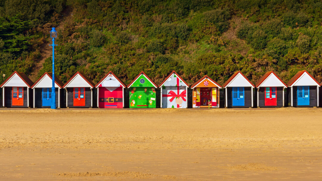 An enchanting 4K wallpaper showcasing a row of festive Christmas beach huts lined along the golden sands of Bournemouth, England. Each hut is uniquely adorned with cheerful holiday decorations, from Santa suits to playful elves, creating a vibrant and joyful seaside Christmas scene.