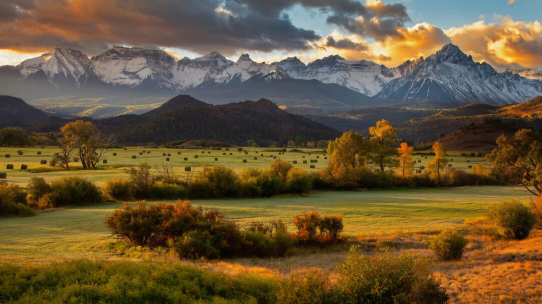 A breathtaking 4K wallpaper captures the scenic Dallas Divide in Southwest Colorado, showcasing a field dotted with autumn hay bales framed by vibrant trees and towering, snow-capped mountains. The dramatic sunset paints the sky with fiery orange and gold hues, casting a warm glow over the landscape and highlighting the rugged textures of the distant peaks.