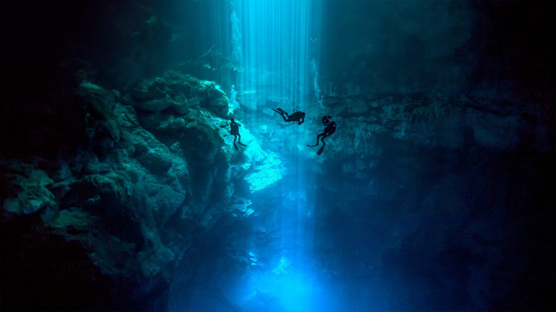 An awe-inspiring 4K wallpaper presents three SCUBA divers exploring the vast, dark depths of El Pit Cenote, part of the Sistema Dos Ojos underwater cave system. Dramatic shafts of sunlight pierce the water, illuminating the cave's rocky formations in stunning shades of turquoise and deep blue, creating a magical, ethereal atmosphere.