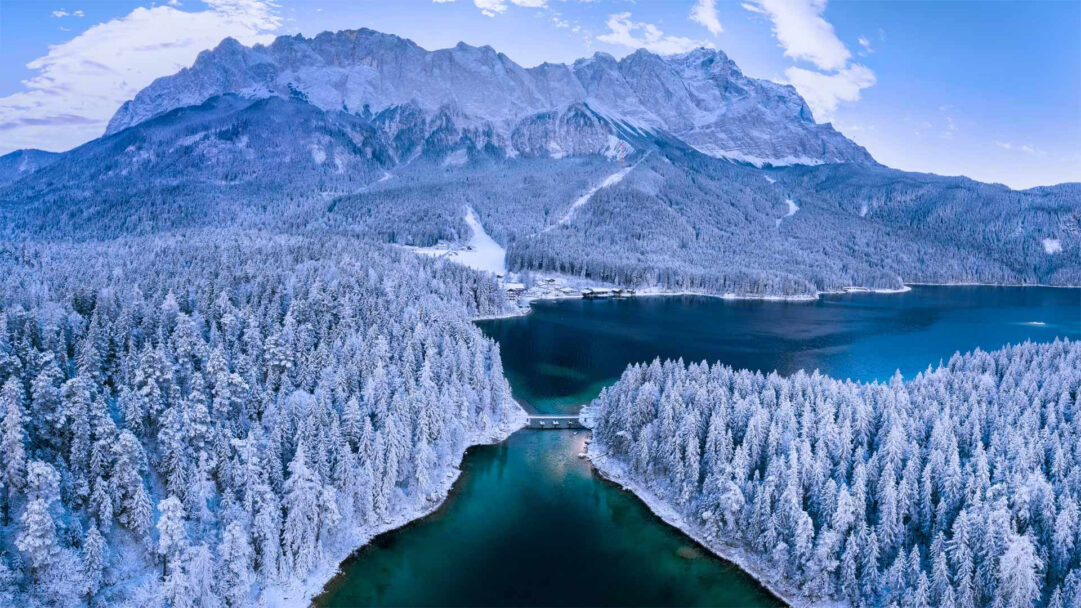 A breathtaking 4K wallpaper capturing the serene Eibsee Lake winter landscape in Bavaria, flanked by a snow-covered forest and the majestic Zugspitze Mountain. The deep, clear turquoise of Eibsee Lake beautifully contrasts with the pristine white of the extensive snow-covered forest and the majestic, icy peaks of the Zugspitze, creating an atmosphere of stunning winter tranquility.