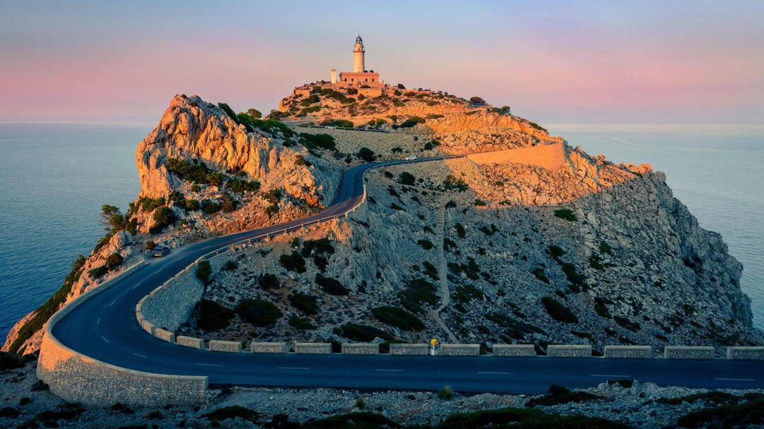 A breathtaking 4K wallpaper featuring the iconic Formentor Lighthouse perched atop a rugged mountain at Cap de Formentor, with a dramatic winding road leading up to it. The golden light of sunset bathes the textured cliffs and lighthouse, contrasting beautifully with the deep blue sea and the soft pastel sky, creating a majestic and serene mood.