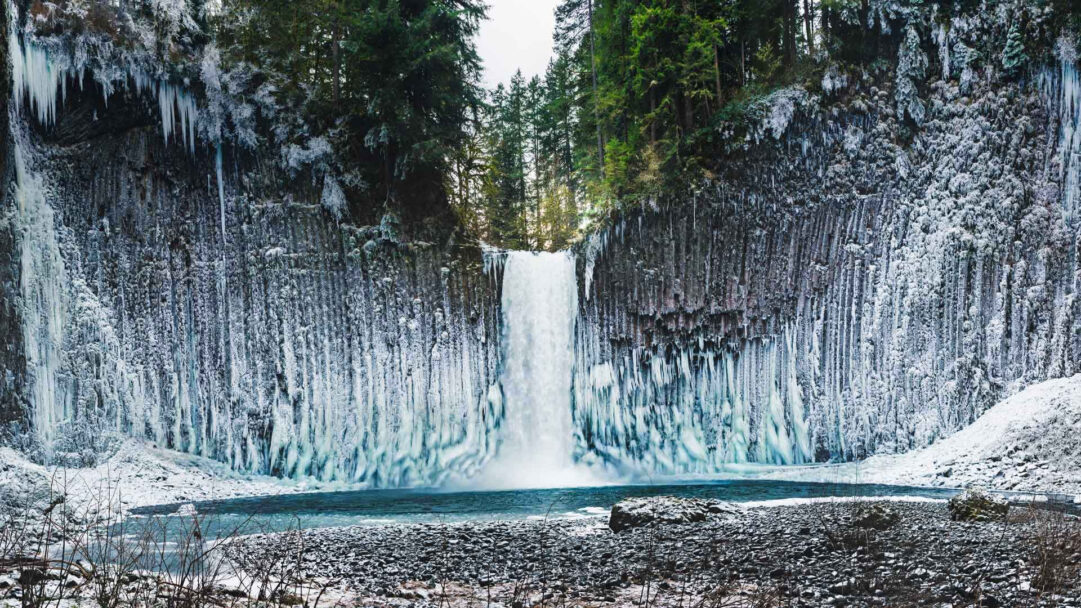A majestic 4K wallpaper showcasing Abiqua Falls in its frozen winter splendor, with a powerful waterfall cascading amidst towering columnar basalt cliffs. Vast sections of the unique basalt columns are dramatically cloaked in brilliant white and blue ice formations, creating a striking contrast with the dark rock and evergreen trees.