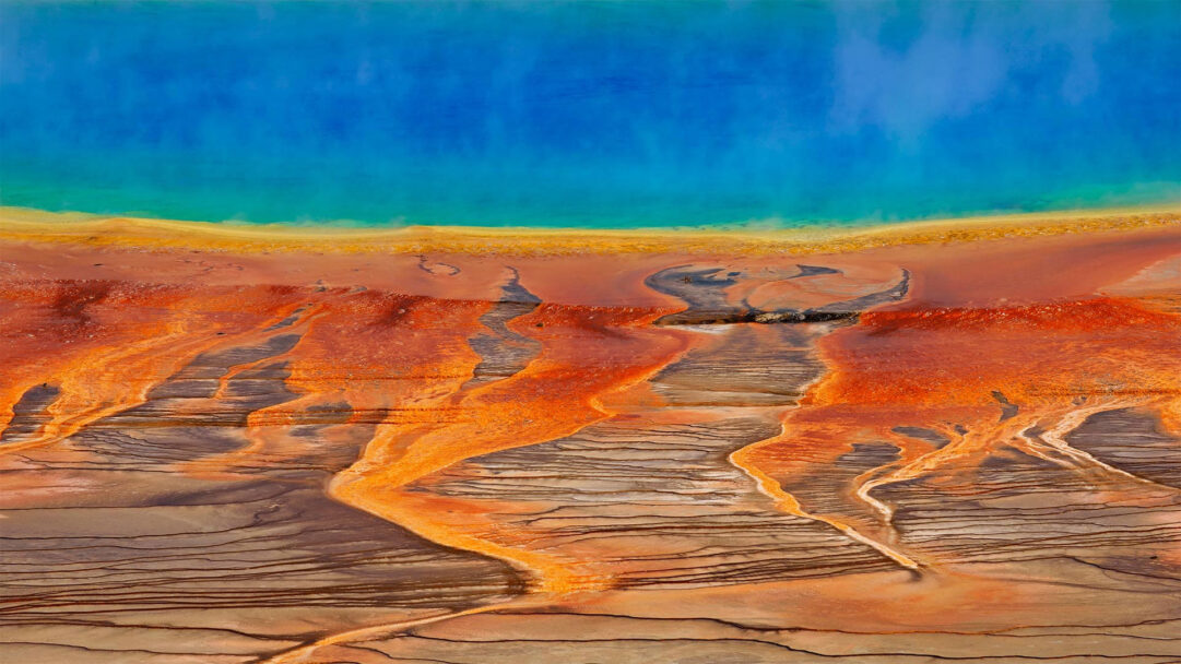 A breathtaking 4K wallpaper capturing the mesmerizing geothermal colors of Grand Prismatic Spring in Yellowstone National Park. Its vivid spectrum transitions from deep blues and greens in the spring's center to stunning fiery oranges and browns across the terraced microbial mats.