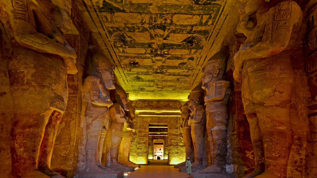 A captivating 4K wallpaper presenting the majestic interior hall of the Great Temple of Ramesses II, where colossal pharaonic statues line both sides, leading the eye into the depths of the ancient structure. The warm, golden illumination bathes the towering figures and ancient hieroglyph-covered ceiling, evoking a powerful sense of awe and timeless grandeur.