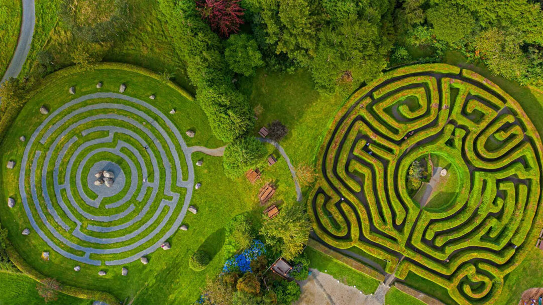 An enchanting 4K wallpaper presents an aerial view of the Greenan Maze in County Wicklow, Ireland, showcasing both a complex hedge labyrinth and a distinct circular stone maze. The vibrant interplay of deep green hedges and light grey stone paths creates a captivating geometric artistry, inviting a sense of wonder and playful exploration.