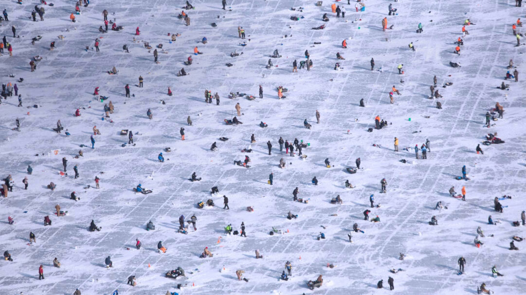 A panoramic 4K wallpaper captures the expansive Gull Lake Brainerd winter scene, densely populated by numerous individuals engaged in ice fishing across its frozen, snow-dusted surface. Brightly dressed figures, scattered widely in a grid-like pattern, create a vibrant mosaic against the vast white expanse of ice, conveying a lively communal spirit despite the frosty conditions.
