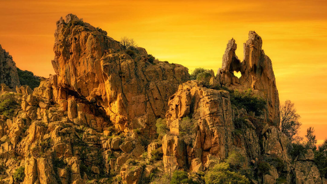 An awe-inspiring 4K wallpaper of a majestic rock formation featuring a distinct heart-shaped hole, set against a stunning sunset sky. The golden-orange hues of the evening sun dramatically illuminate the rugged textures of the ancient rocks, creating a warm and romantic ambiance.
