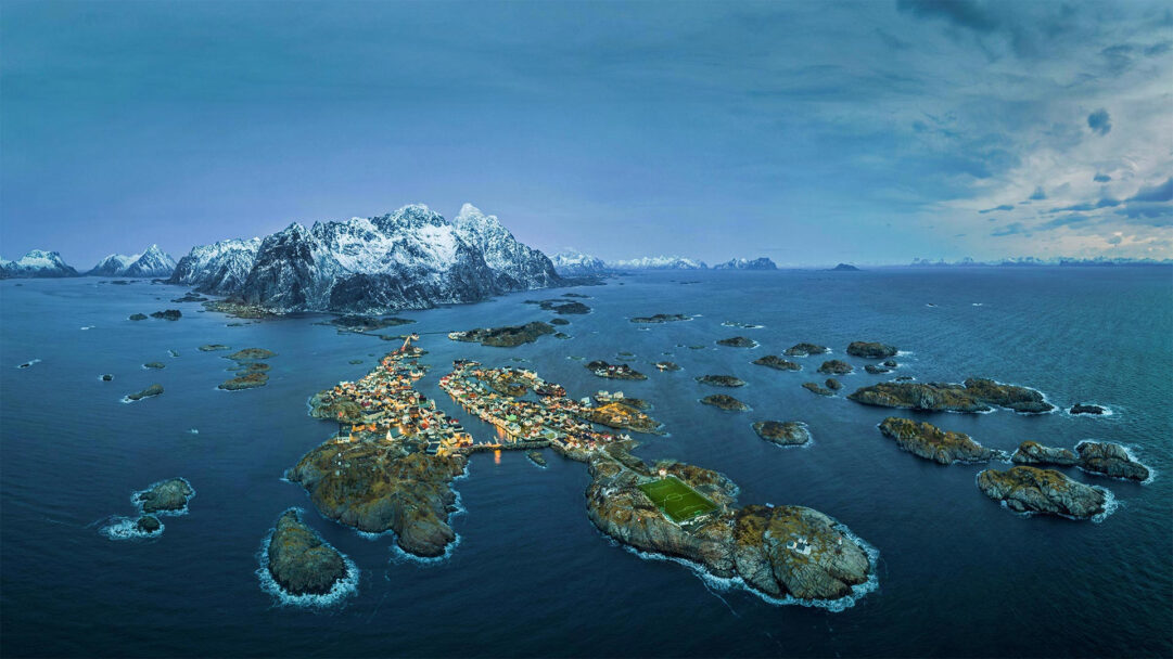 An expansive 4K wallpaper depicts the iconic Henningsvær Fishing Village and its unique stadium nestled on rocky islets in Norway's deep blue waters. The village's warm lights glow softly against the dramatic backdrop of snow-capped mountains and a dusky sky, while the vibrant green soccer pitch stands out amidst the rugged maritime landscape.