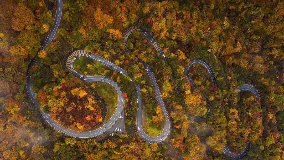 An enthralling 4K wallpaper capturing the intensely winding Irohazaka Road in Nikko, Japan, as it snakes through a dense autumn forest. Its intricate curves are spectacularly framed by a rich tapestry of red, orange, and golden trees, with distant hints of mist adding depth to the vibrant seasonal display.