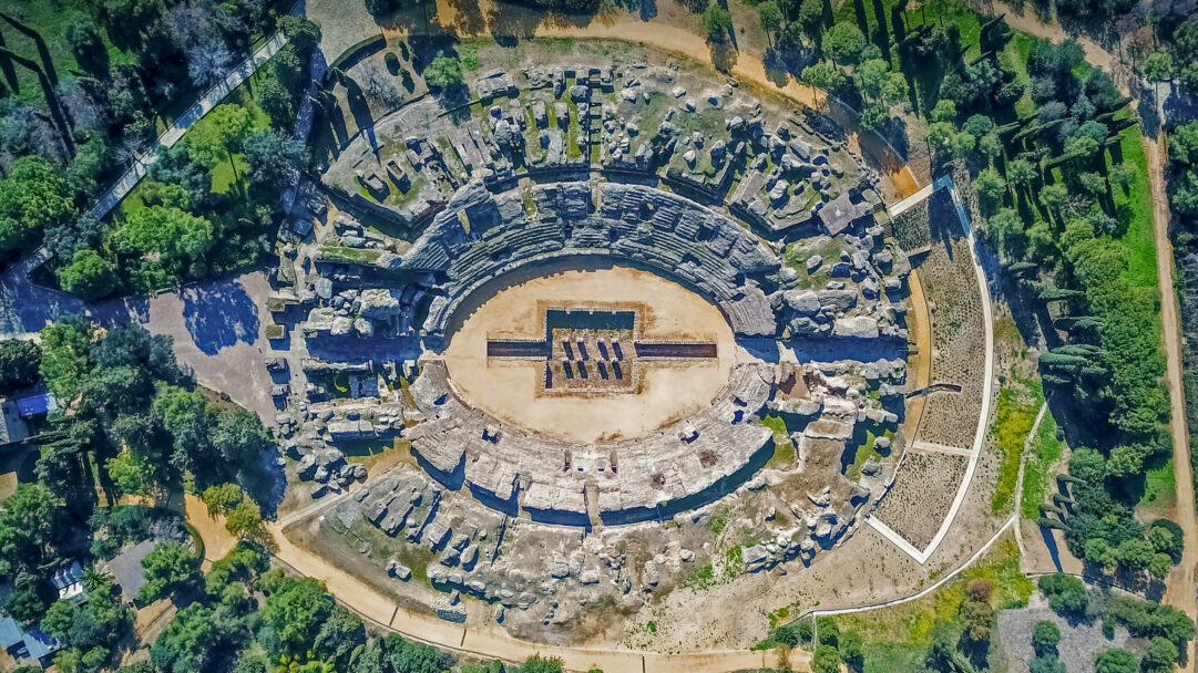 An awe-inspiring 4K wallpaper presents an aerial view of the ancient Italica Roman Amphitheater in Seville, Spain, surrounded by lush green landscapes. The remarkably preserved oval structure, with its sandy arena and fragmented stone seating, is dramatically illuminated by sunlight, casting sharp shadows that emphasize its historical depth amidst the vibrant trees.