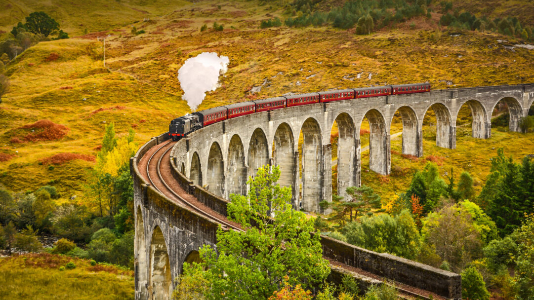 A captivating 4K wallpaper showcasing the iconic Jacobite Steam Train as it majestically crosses the curving Glenfinnan Viaduct amidst a vibrant Scottish landscape. The train's dark silhouette and plume of white steam dramatically stand out against the rich tapestry of golden-yellow and deep green hills, capturing the enchanting autumnal beauty of Scotland.