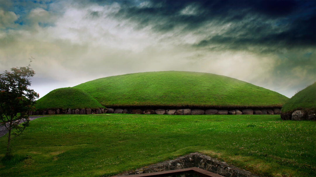 A majestic 4K wallpaper captures the ancient Knowth Burial Mound in Ireland's historic Boyne Valley, standing as a prominent grassy hill amidst a lush landscape. The vibrant green of the mound's meticulously maintained turf sharply contrasts with the dramatic, brooding sky overhead, evoking a sense of timeless mystery.