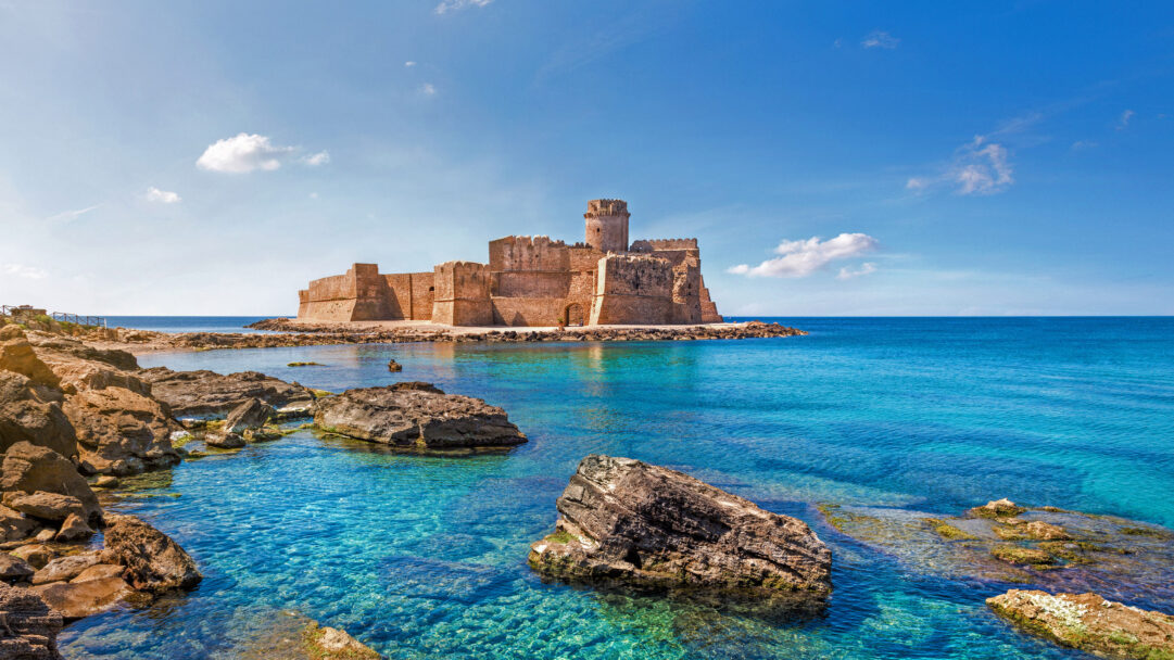 A captivating 4K wallpaper showcases Le Castella Castle in Capo Rizzuto, Italy, majestically situated on a rocky islet in a brilliant blue sea. The crystal-clear turquoise waters reveal submerged rocks near the shore, beautifully contrasting with the warm, ancient stone of the fortress under a bright, cloud-dotted sky.