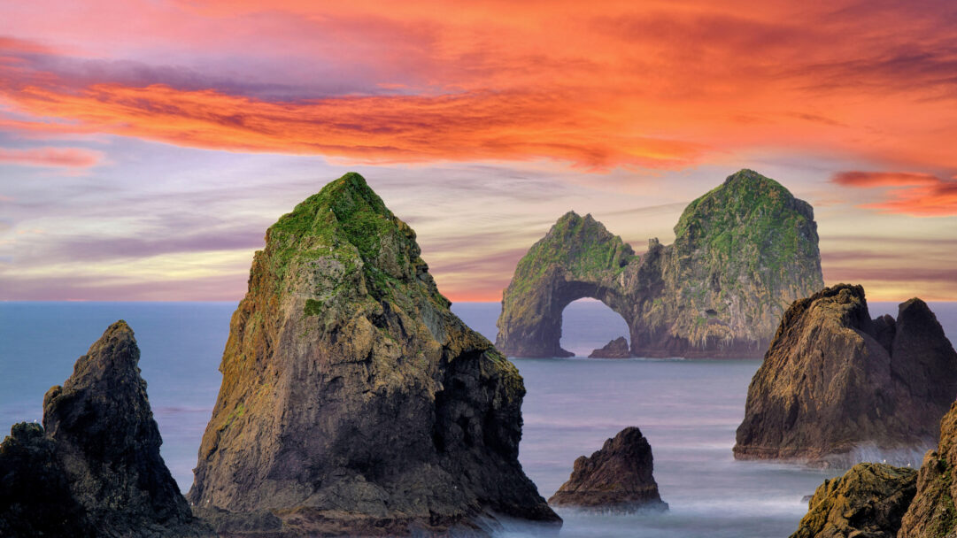 This breathtaking 4K wallpaper presents Mack Arch Rock and other rugged sea stacks rising from the Pacific Ocean along the Southern Oregon Coast at sunrise. The fiery orange and pink hues of the sunrise illuminate the dramatic sky, casting a mystical glow over the ancient, moss-covered rock formations and the gentle, misty ocean waves.