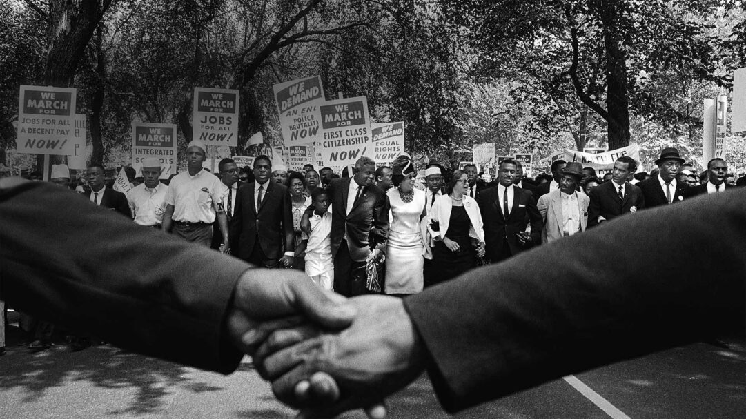 A historic 4K wallpaper capturing the iconic March on Washington, depicting a determined crowd of civil rights activists moving forward under a canopy of trees. In the foreground, a prominent handshake symbolizes the profound unity and shared purpose of the movement, rendered in timeless black and white.