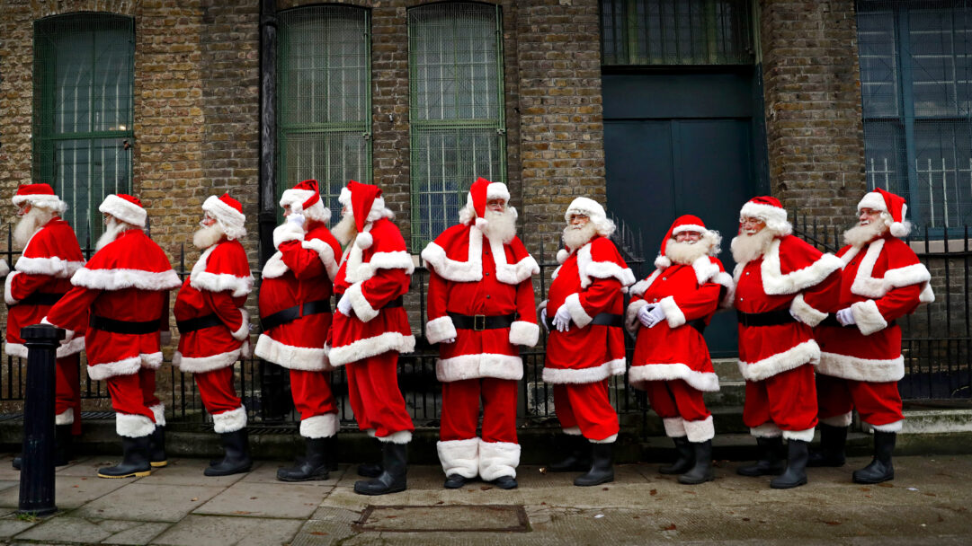 An enchanting 4K wallpaper captures Ministry of Fun Santa School London performers in their full festive attire, lined up neatly on a city sidewalk. Their bright red suits and white beards stand out vividly against the subdued, textured brick wall, creating a cheerful Christmas atmosphere.
