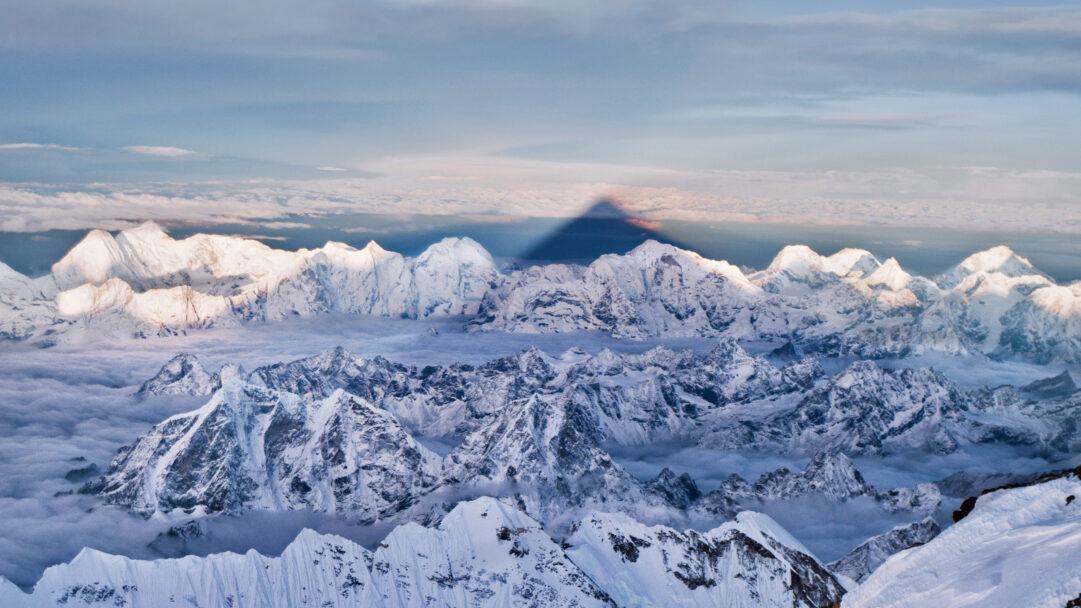 A majestic 4K wallpaper of Mount Everest's colossal triangular shadow cast across a vast panorama of sunlit snow mountains and a swirling sea of clouds in the Western Himalayas. The stark, elongated shadow dramatically bisects the sky, contrasting with the vibrant orange hues on the distant cloud line and the frosty white peaks, conveying a profound sense of scale and natural grandeur.