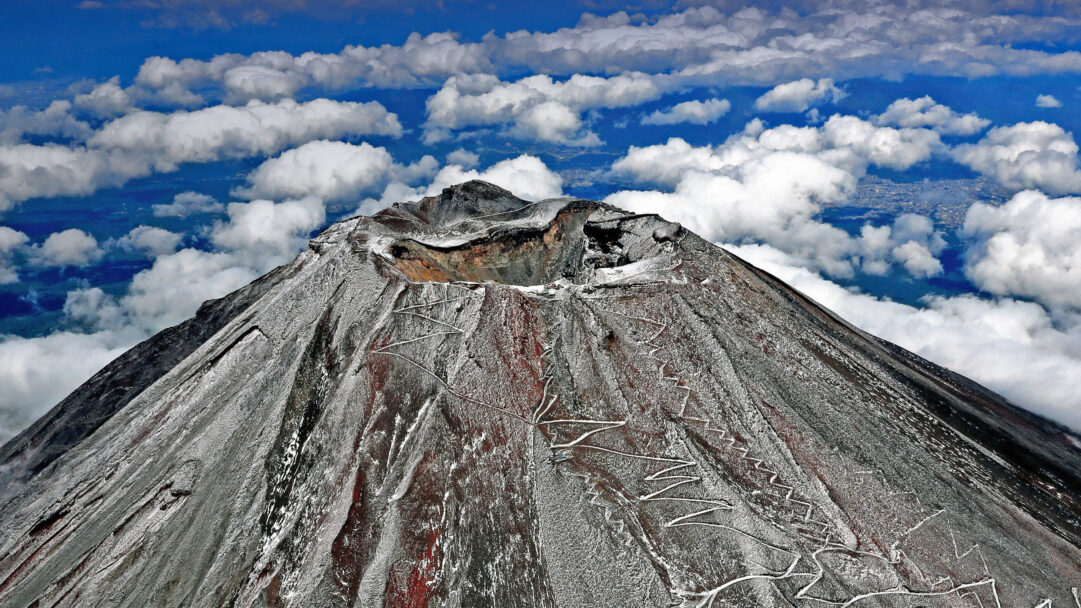 A majestic 4K wallpaper presents Mount Fuji's snowcapped summit rising high above a vast expanse of clouds in Japan. The rugged, partially snow-dusted slopes, revealing hints of reddish rock, dramatically pierce through the soft white cloud blanket, set against a deep blue sky.
