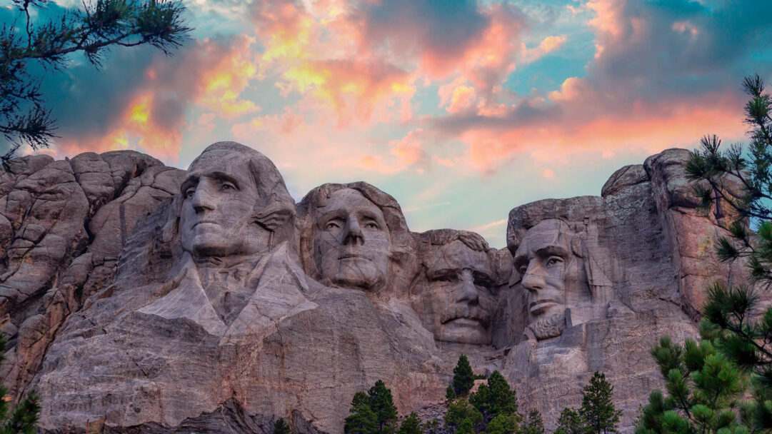 A breathtaking 4K wallpaper showcasing the iconic Mount Rushmore National Memorial in South Dakota at sunset. The colossal stone carvings of the four presidents are dramatically silhouetted against a vibrant sky painted with brilliant pink, orange, and blue clouds, evoking a majestic and patriotic mood.