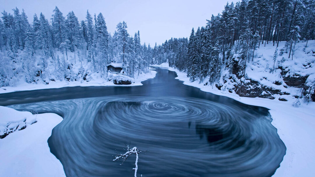 A captivating 4K wallpaper showcasing the Myllykoski Scenic Area, a serene winter landscape where a river carves its path through a dense, snow-laden forest. The dark waters feature a prominent, mesmerizing whirlpool effect in the foreground, contrasting beautifully with the pristine white snow and muted blue sky.