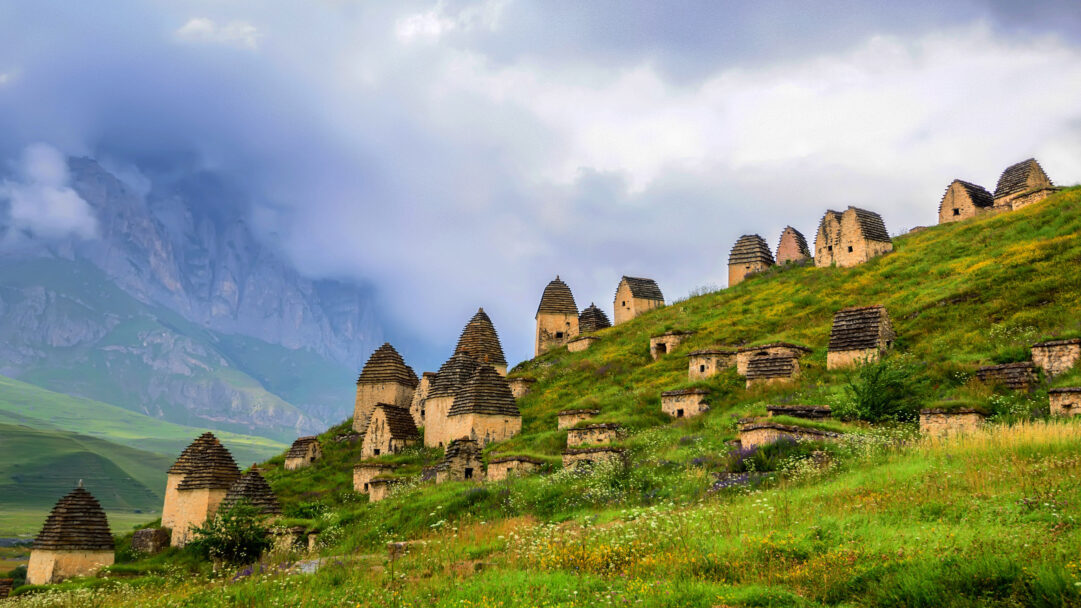 An ethereal 4K wallpaper captures the ancient Necropolis of Dargavs, featuring numerous cone-roofed tombs clustered on a verdant, wildflower-covered hillside in North Ossetia, Russia. Dramatic clouds enshroud the towering mountain peaks behind, casting a mystical atmosphere over the unique ancient structures and the vibrant hillside blooming with wildflowers.