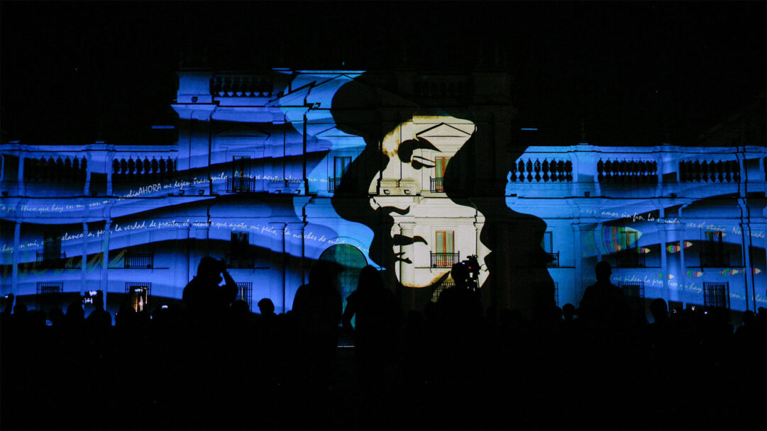 A poetic 4K wallpaper illuminating a grand building at night with a large projected portrait of Pablo Neruda. His iconic profile is rendered in warm tones, contrasting with the cool blue light and flowing white script across the building, as an audience gathers below in silhouette.