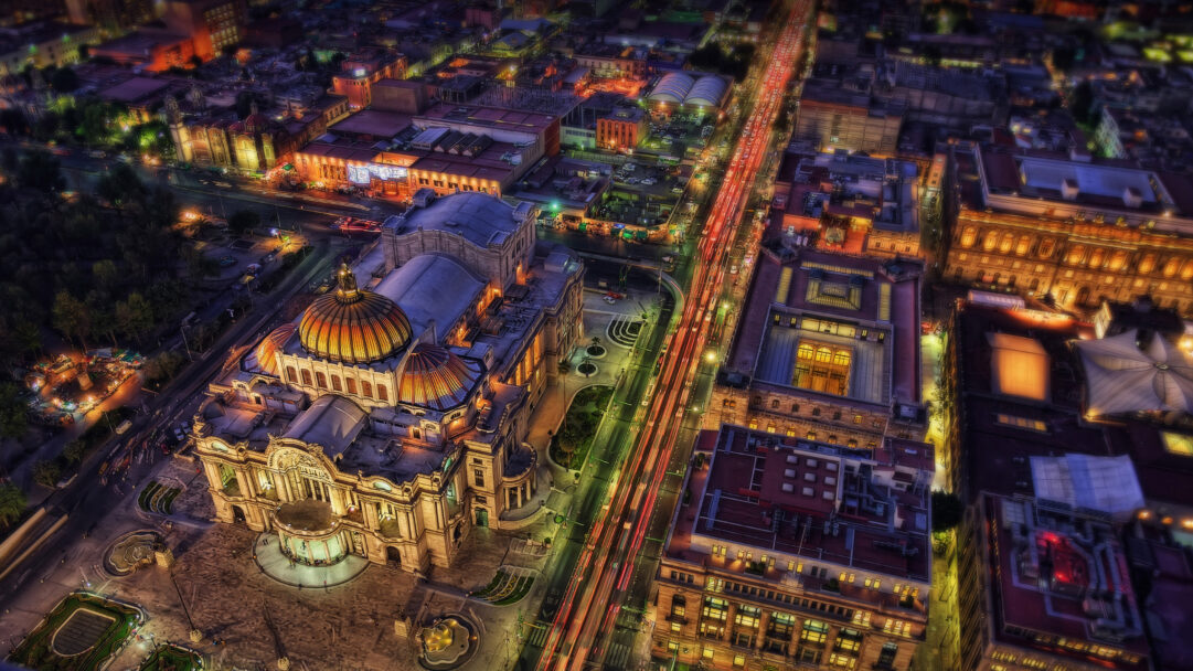 An enchanting 4K wallpaper captures the Palacio de Bellas Artes in Mexico City, seen from an aerial perspective at night, surrounded by the illuminated cityscape. Its distinctive golden dome and detailed facade glow warmly amidst the blur of red and white vehicle light trails on the bustling streets below, creating a vibrant, dynamic urban scene.